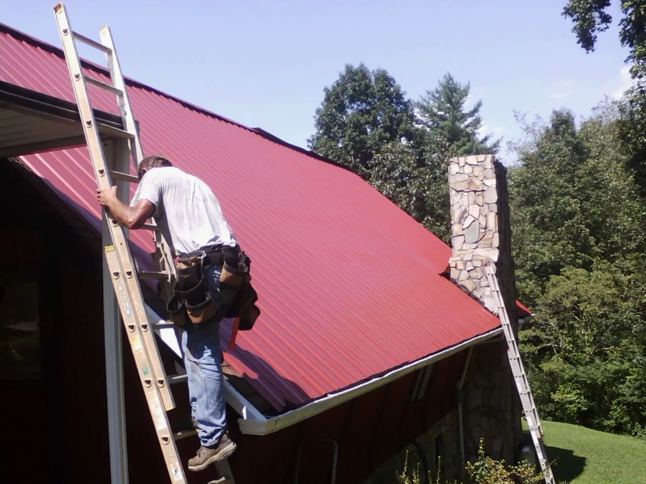 Roofer on a ladder beside a red metal roof and stone chimney — Soffit & Fascia Repair by Roofing Brothers in Trivoli